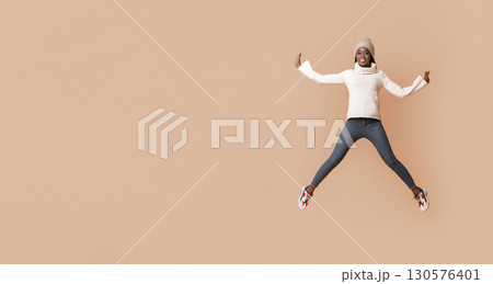 Emotional african woman in winter hat jumping up, spreading legs and hands widely, beige studio background 130576401