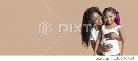 Mother's love. Happy african american mom cuddling her cute little daughter from the back, yellow background with free space Mother's love. Happy african american mom cuddling her cute little daughter from the back, yellow background with free space 130576414