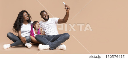 Selfie with family. Cheerful black father taking photo with his daughter and wife, posing together on floor over yellow studio background. Selfie with family. Cheerful black father taking photo with his daughter and wife, posing together on floor over yellow studio background. 130576415