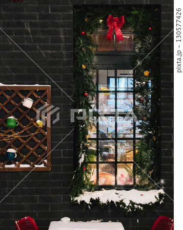 Christmas garland with red bow, baubles and dried oranges adorns window of cozy cafe during gentle snowfall, creating festive and inviting atmosphere. Festive window decor, red ribboned garland Christmas garland with red bow, baubles and dried oranges adorns window of cozy cafe during gentle snowfall, creating festive and inviting atmosphere. Festive window decor, red ribboned garland 130577426