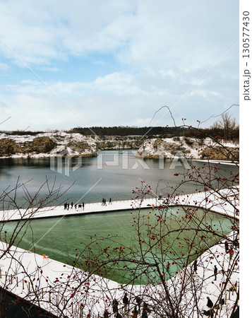 Tourists are walking near an artificial body of water created in former quarry, during winter, with snow covering ground and red berries on branches. Tourists walking near flooded limestone quarry Tourists are walking near an artificial body of water created in former quarry, during winter, with snow covering ground and red berries on branches. Tourists walking near flooded limestone quarry 130577430