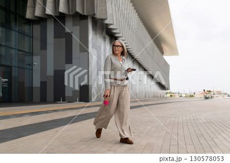 Woman walking in casual attire along a modern building in a coastal city at midday 130578053