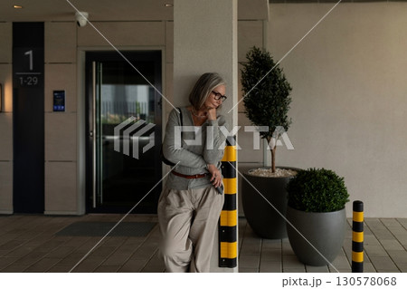 Senior woman leaning against a post while waiting outside a modern building during daylight hours Senior woman leaning against a post while waiting outside a modern building during daylight hours 130578068
