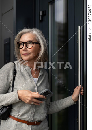 Woman with glasses steps outside a building while checking her smartphone on a sunny day 130578070
