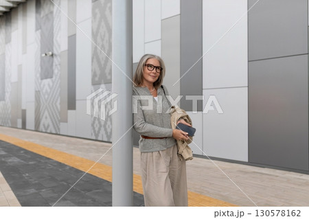Confident woman stands by modern urban wall while holding phone and jacket in late afternoon 130578162