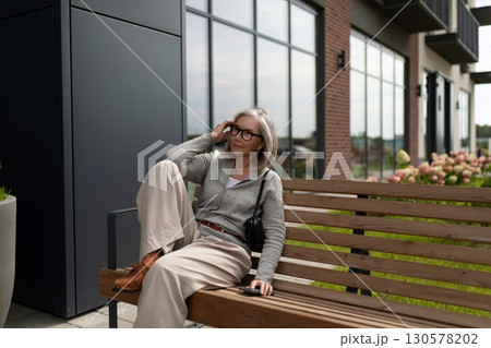 Mature woman relaxes on a bench outside a modern building during a cloudy day 130578202