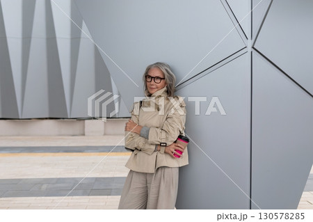 Stylish older woman leans against modern architecture while enjoying a coffee in an urban setting Stylish older woman leans against modern architecture while enjoying a coffee in an urban setting 130578285