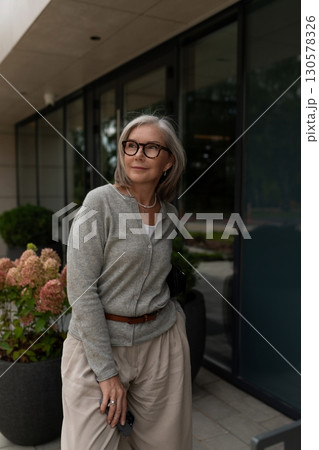 Elderly woman in stylish outfit enjoying a sunny day outside modern building with plants 130578326