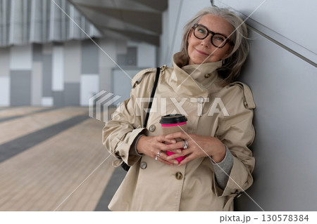 Stylish woman enjoying coffee while leaning against a wall in an urban environment during the day 130578384