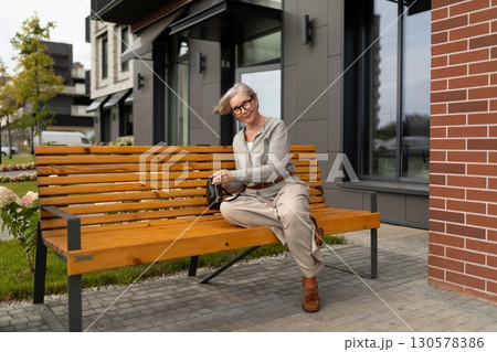 Elderly woman sits on wooden bench outside modern building enjoying peaceful afternoon in a suburban 130578386