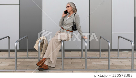 Older woman casually talking on mobile phone while sitting on railings outside modern building Older woman casually talking on mobile phone while sitting on railings outside modern building 130578395