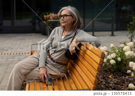 Elderly woman relaxing on a wooden bench in a modern garden area during a sunny day in an urban Elderly woman relaxing on a wooden bench in a modern garden area during a sunny day in an urban 130578413