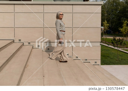 Elderly woman walks confidently down steps in a modern outdoor space during a sunny afternoon stroll 130578447