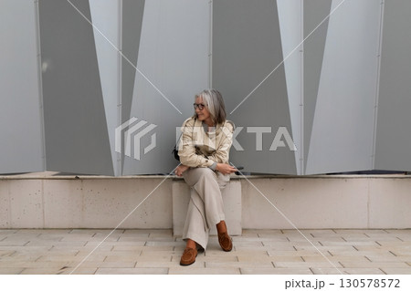 Woman sitting casually on a bench against a modern geometric wall in an urban setting Woman sitting casually on a bench against a modern geometric wall in an urban setting 130578572