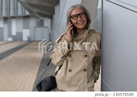 Woman in a beige coat talking on the phone while standing near a modern building in the city during 130578580