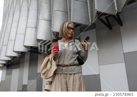 Stylish woman checking her phone while holding coffee outside a modern building in the city during 130578965