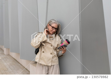 Older woman in beige coat holding coffee cup while checking phone against modern gray wall 130578982