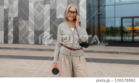 Senior woman talks on phone outside modern building while holding coffee in urban environment Senior woman talks on phone outside modern building while holding coffee in urban environment 130578992