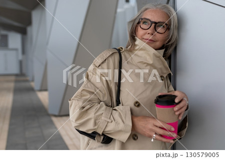 Stylish woman in neutral trench coat enjoying coffee outside modern building on a sunny day 130579005
