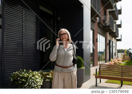 Mature woman checking her phone while standing outside a modern building in a serene urban setting Mature woman checking her phone while standing outside a modern building in a serene urban setting 130579098