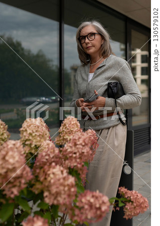Stylish woman stands near blooming flowers outside a modern building on a cloudy day 130579102