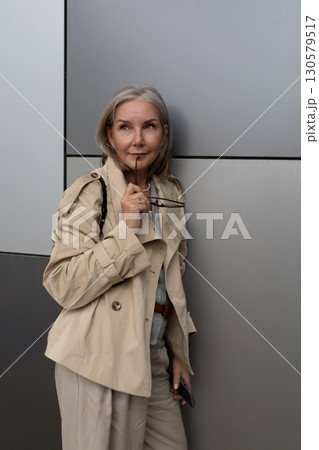 Mature woman in stylish trench coat poses thoughtfully against modern architecture in a vibrant 130579517