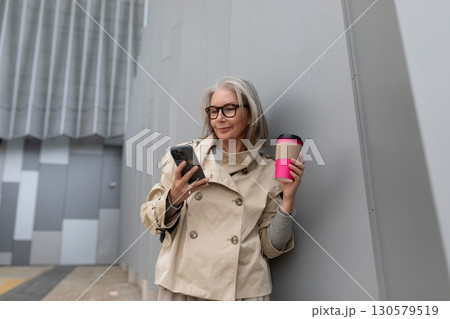 Woman in stylish trench coat checks her smartphone while enjoying a beverage outside a modern 130579519