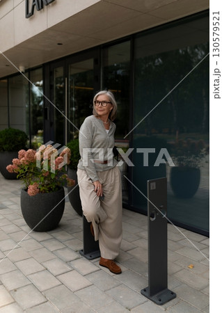 Elderly woman in stylish outfit stands beside potted flowers outside modern building during daytime 130579521