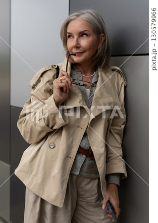 Elegant woman in stylish trench coat poses near modern building in urban setting during sunny day Elegant woman in stylish trench coat poses near modern building in urban setting during sunny day 130579966