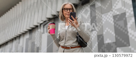 Woman stands in front of modern building holding coffee cup while checking her phone in urban Woman stands in front of modern building holding coffee cup while checking her phone in urban 130579972