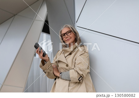 Senior woman in glasses holding a smartphone while leaning against a modern architectural structure Senior woman in glasses holding a smartphone while leaning against a modern architectural structure 130580082