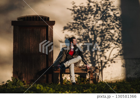 Lonely young man sitting on bench in front of old house, looking at camera. 130581366