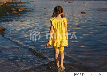 Little girl walking along seashore in yellow dress on sunny day, view from back Little girl walking along seashore in yellow dress on sunny day, view from back 130581699