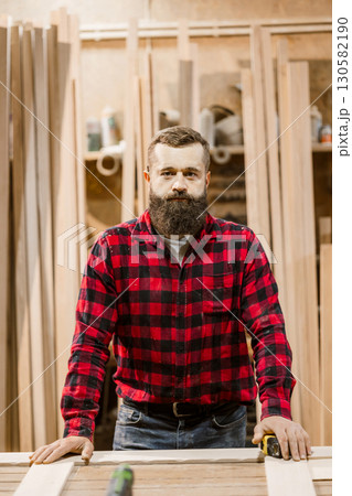 Craftsman in a woodworking shop wearing a plaid shirt, focused on his task amid wooden materials and tools 130582190