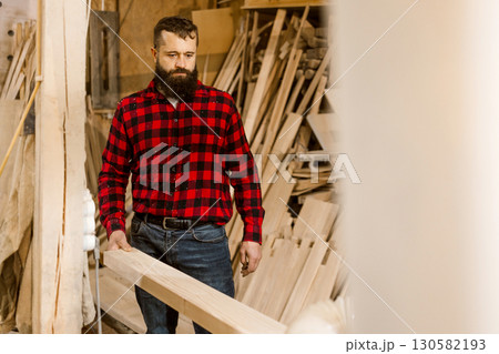 Woodworker carefully handles lumber in a workshop surrounded by tools and timber at daytime Woodworker carefully handles lumber in a workshop surrounded by tools and timber at daytime 130582193