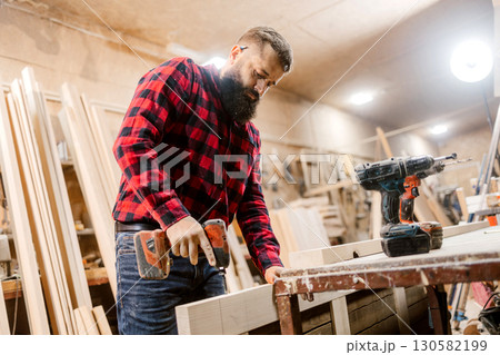 Skilled carpenter working in a workshop using a power drill to build wooden furniture 130582199
