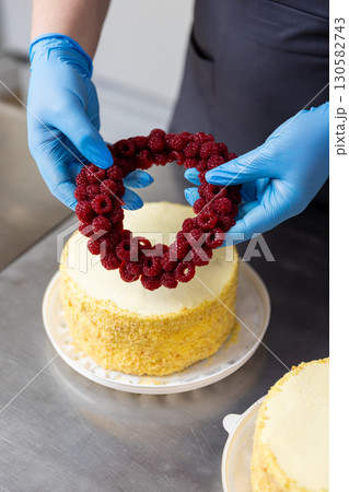 Chef decorating cake with fresh raspberry wreath on top in professional bakery kitchen 130582743