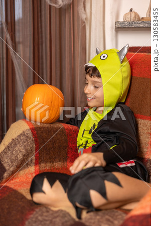Smiling child dressed in a playful halloween monster costume, sitting on a cozy sofa with a blanket and joyfully holding a pumpkin at home, surrounded by festive decorations Smiling child dressed in a playful halloween monster costume, sitting on a cozy sofa with a blanket and joyfully holding a pumpkin at home, surrounded by festive decorations 130583455