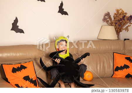 Smiling boy in a monster costume holding a plush spider sits on a bat decorated sofa with a pumpkin, ready for halloween celebrations Smiling boy in a monster costume holding a plush spider sits on a bat decorated sofa with a pumpkin, ready for halloween celebrations 130583978