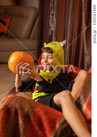 Happy child wearing a playful monster costume, holding a bright pumpkin while sitting comfortably on a sofa in a festively decorated living room, celebrating halloween at home 130583980