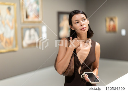 Woman with phone in hands in museum hall, view painting exhibition Woman with phone in hands in museum hall, view painting exhibition 130584290