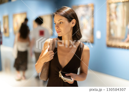 Woman with phone in hands in museum hall, view painting exhibition Woman with phone in hands in museum hall, view painting exhibition 130584307