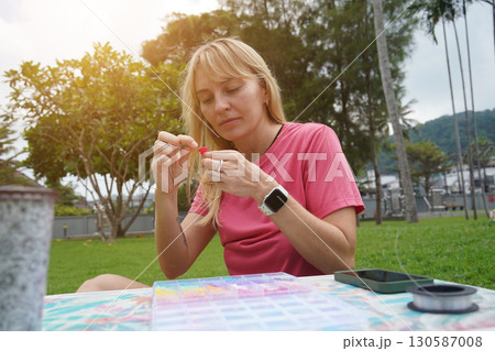 Young woman beading colorful bracelets outdoors in a park 130587008