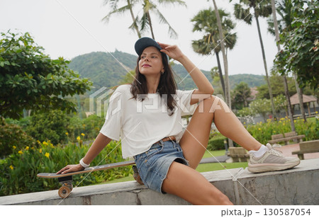 A young woman sits comfortably on a low wall at the park holding a skateboard in her hands 130587054