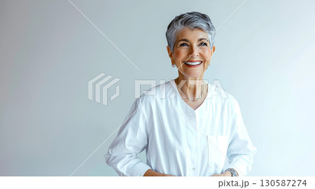Smiling elderly woman in white shirt standing against light background Smiling elderly woman in white shirt standing against light background 130587274
