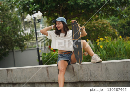 A young woman sits comfortably on a low wall at the park holding a skateboard in her hands A young woman sits comfortably on a low wall at the park holding a skateboard in her hands 130587465