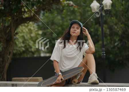 A young woman sits comfortably on a low wall at the park holding a skateboard in her hands A young woman sits comfortably on a low wall at the park holding a skateboard in her hands 130587469