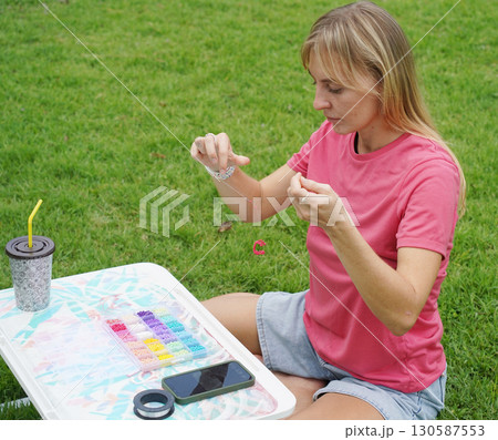 Young woman beading colorful bracelets outdoors in a park Young woman beading colorful bracelets outdoors in a park 130587553