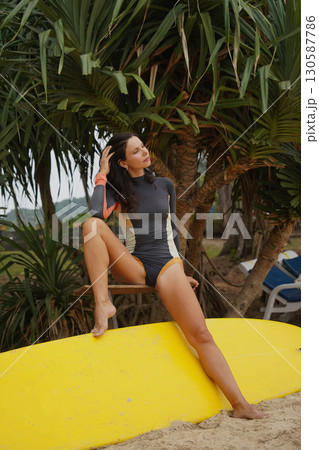 Woman in stylish wetsuit posing confidently with surfboard on beach Woman in stylish wetsuit posing confidently with surfboard on beach 130587786
