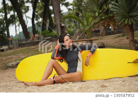 Woman in stylish wetsuit posing confidently with surfboard on beach Woman in stylish wetsuit posing confidently with surfboard on beach 130587952